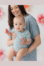 A smiling woman holding a baby wearing matching light blue patterned baby bodysuits and a blue floral dress, set against a soft white background with blurred pink flowers. 