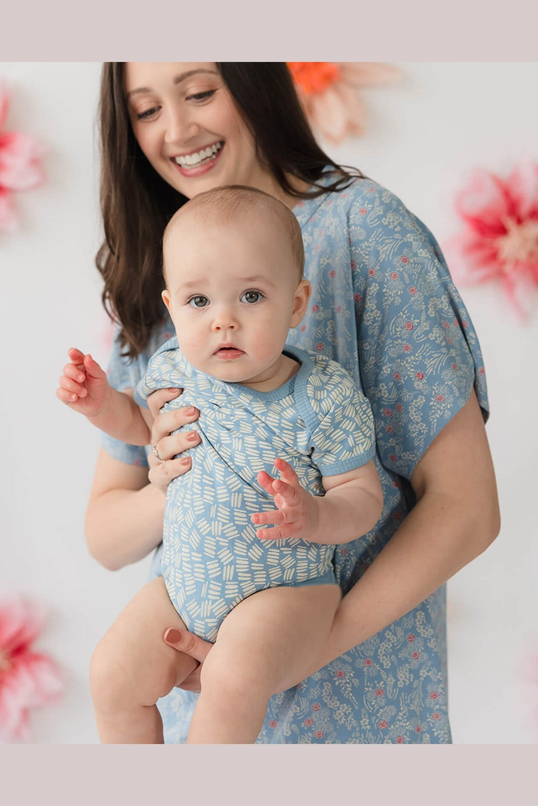 A smiling woman holding a baby wearing matching light blue patterned baby bodysuits and a blue floral dress, set against a soft white background with blurred pink flowers. 
