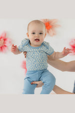 A baby wearing a light blue patterned short-sleeve top and matching blue pants, being held up by an adult's hands against a white background with soft pink floral decorations. 