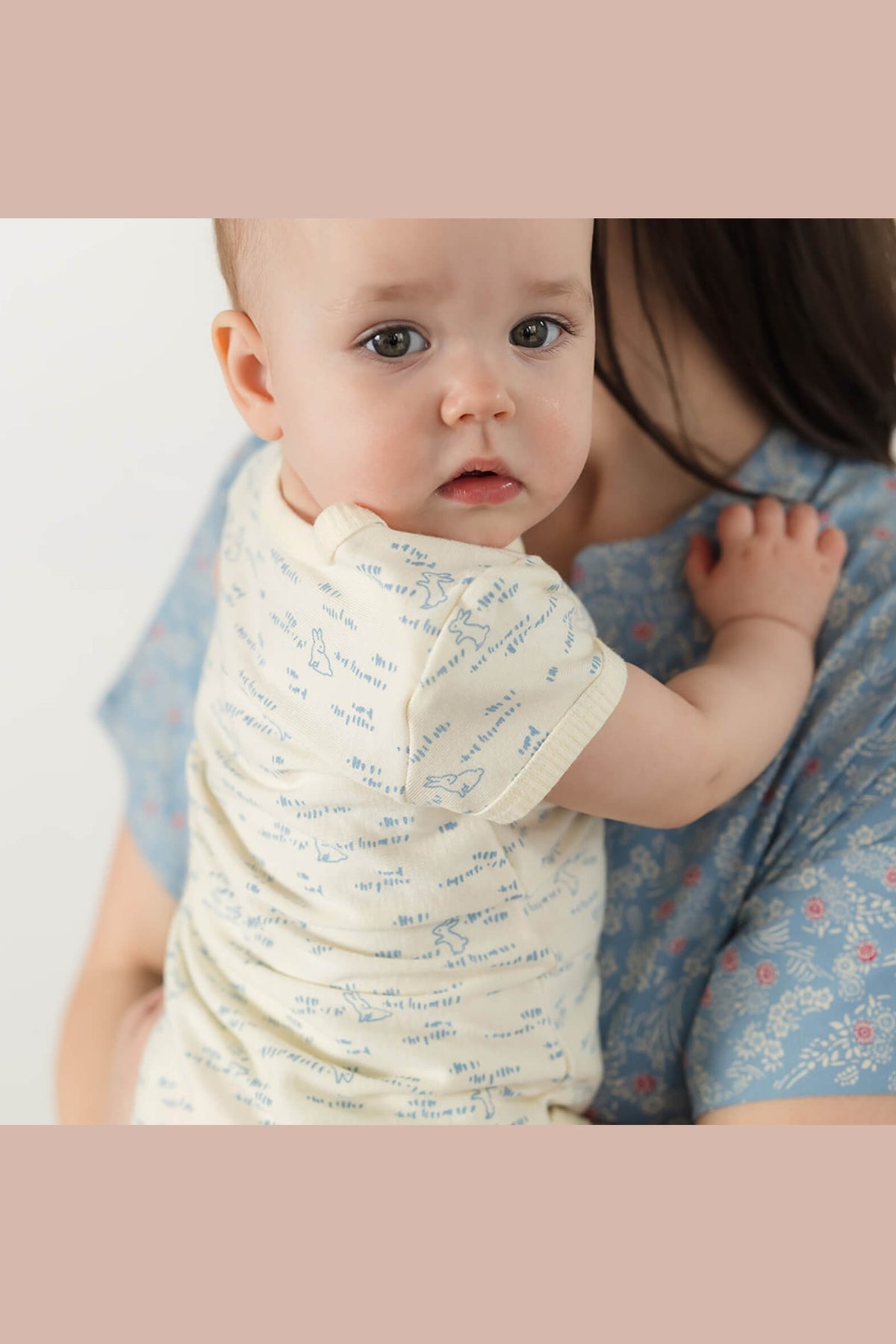 A baby wearing a cream short-sleeve bodysuit with a light blue script pattern, held by an adult wearing a blue floral shirt. 