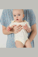 A baby wearing a cream-colored bodysuit with a light blue pattern, held by an adult wearing a blue floral shirt. The photo is framed with gray bars on the top and bottom. 