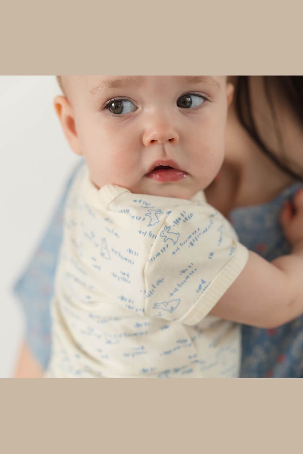 A close-up lifestyle shot of a baby wearing a cream-colored short-sleeve romper with blue handwritten text and star patterns, held by an adult. 