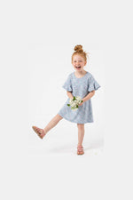 A young girl with red hair in a bun is wearing a light blue short-sleeve dress with a small floral print and holding a bouquet of white flowers. She is wearing pink sandals and posing on a solid white studio background.