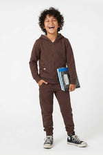 A young boy with curly dark hair is smiling and laughing while wearing a matching brown zip-up hoodie and jogger sweatpants set. He is holding a blue notebook and standing against a clean white studio background. 
