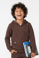 A smiling young boy with curly dark hair wearing a brown zip-up hoodie and matching brown sweatpants, holding a blue and black patterned notebook against a plain white background. 