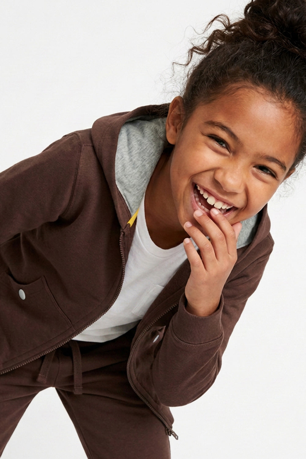 A young girl smiling and laughing while wearing a dark brown hooded sweatshirt with a gray inner hood lining, a white t-shirt, and matching brown sweatpants against a plain white background. 