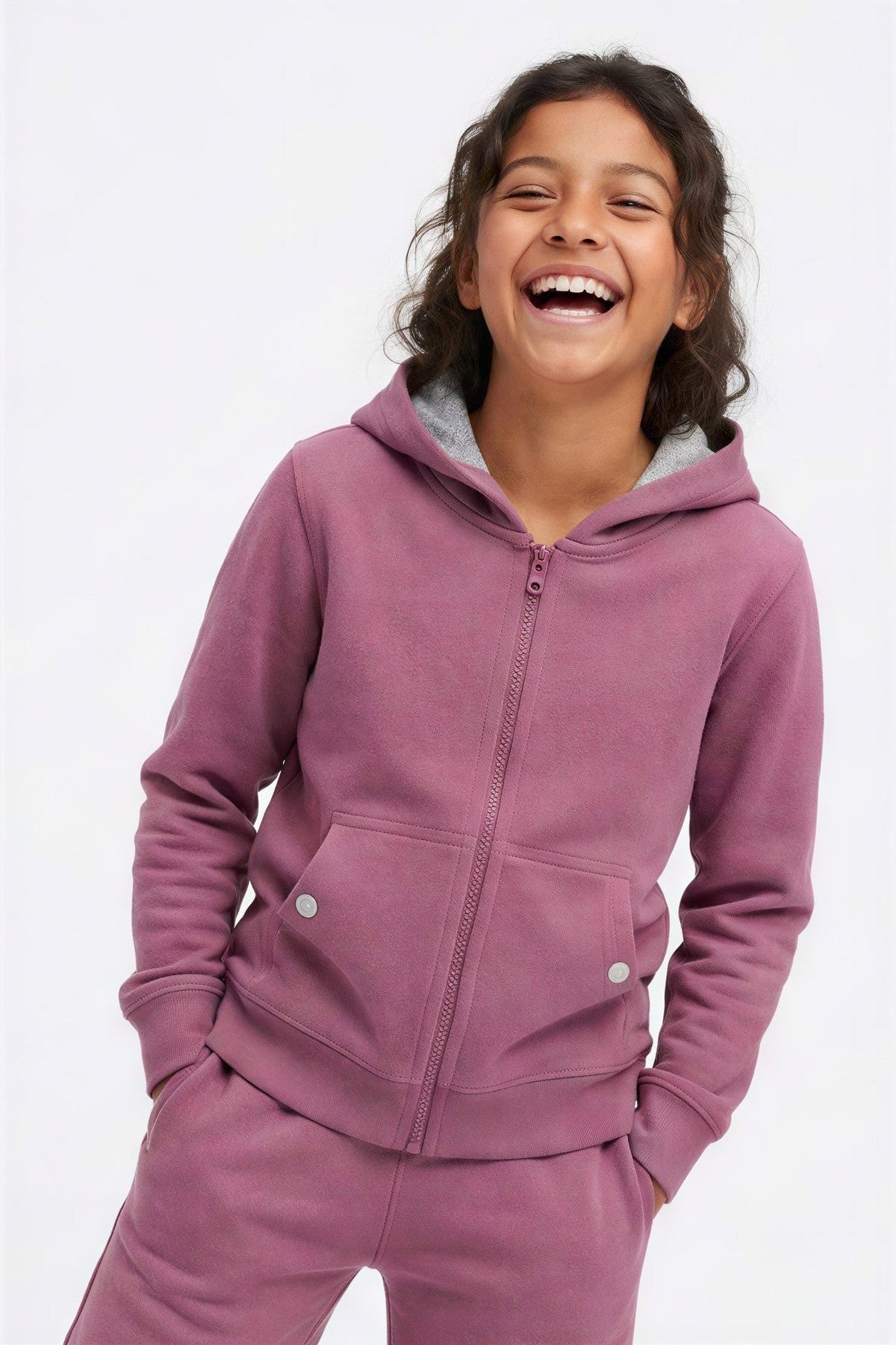A young girl smiling and laughing while wearing a dusty mauve zip-up hoodie with a gray hood lining and matching sweatpants, photographed against a clean white background. 