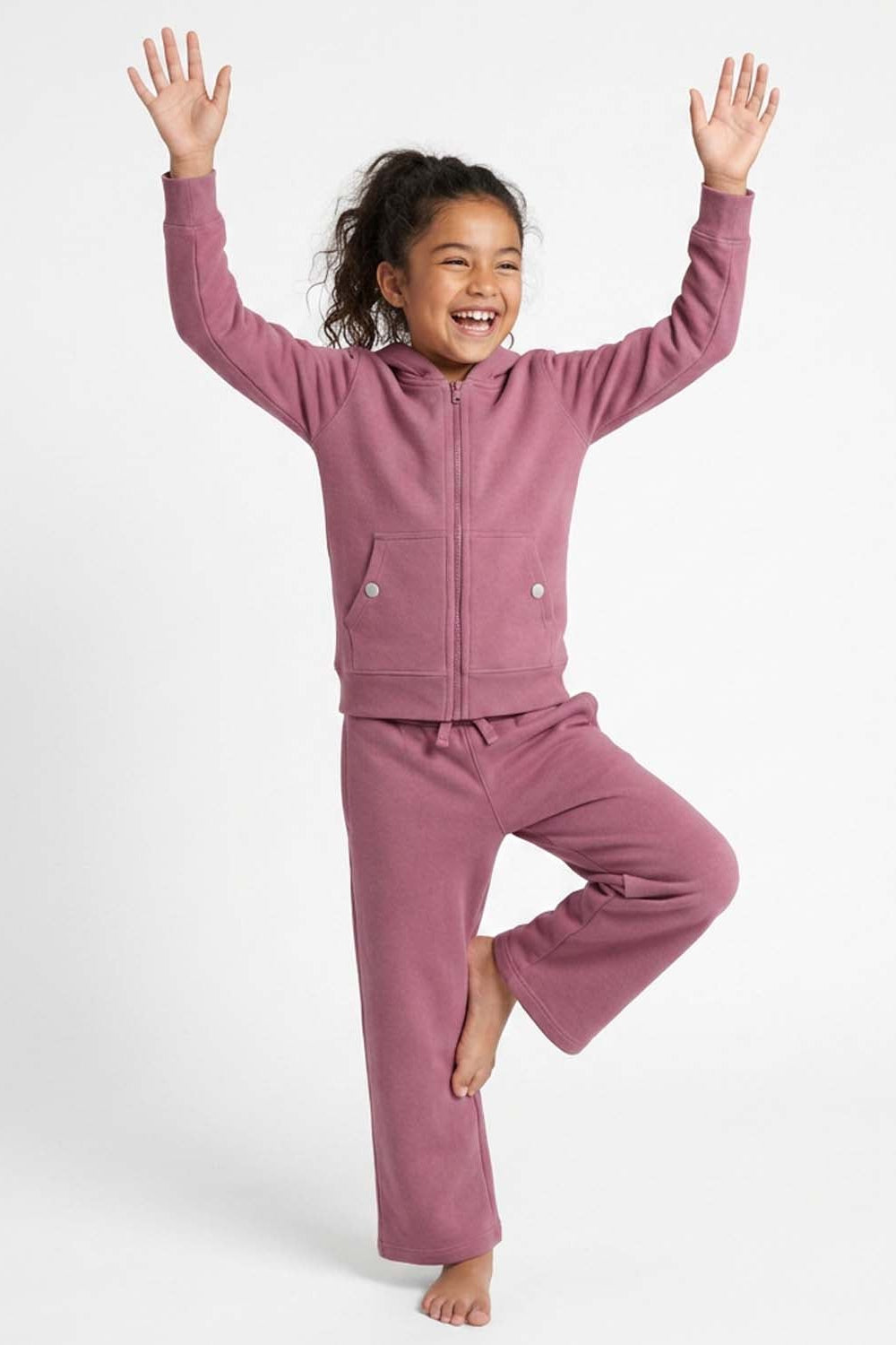 A young girl smiling and posing in a matching dusty rose fleece zip-up hoodie and sweatpants set against a clean white studio background. 