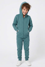 A young boy smiling while wearing a matching teal green zip-up hoodie and jogger sweatpants set, paired with white sneakers, against a plain white studio background. 