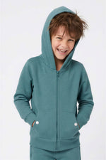 A young boy smiling while wearing a teal zip-up hoodie with a matching pair of pants, photographed against a clean white studio background. 