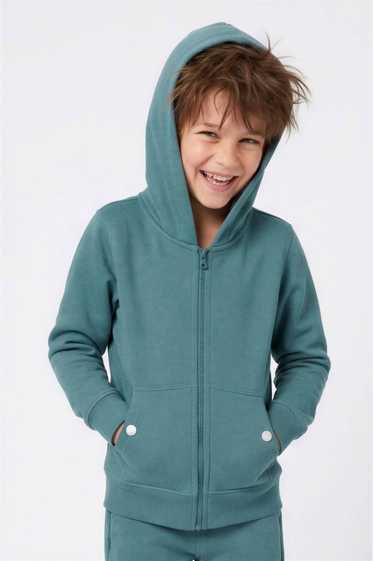 A young boy smiling while wearing a teal zip-up hoodie with a matching pair of pants, photographed against a clean white studio background. 