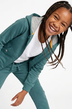 A young girl with long braids smiling and laughing while wearing a teal green zip-up hoodie and matching sweatpants over a white t-shirt, photographed against a clean white background. 
