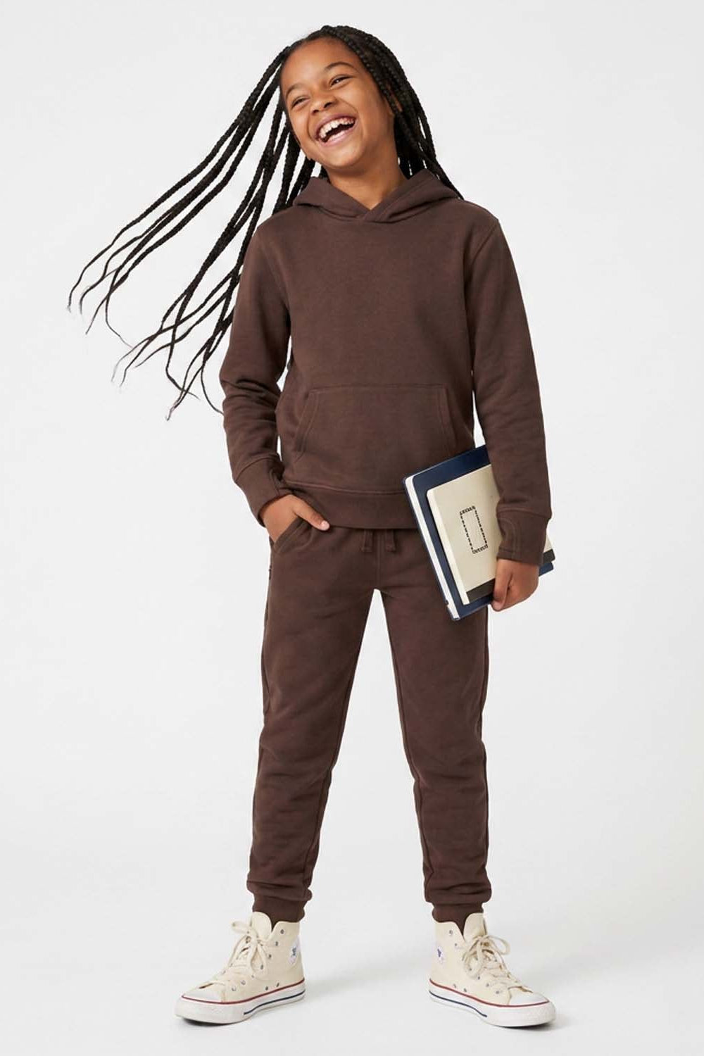 A young girl with long black dreadlocks is smiling and laughing while modeling a matching brown sweatshirt and jogger pants set. She is holding a notebook and wearing off-white high-top sneakers, standing against a plain white studio background. 