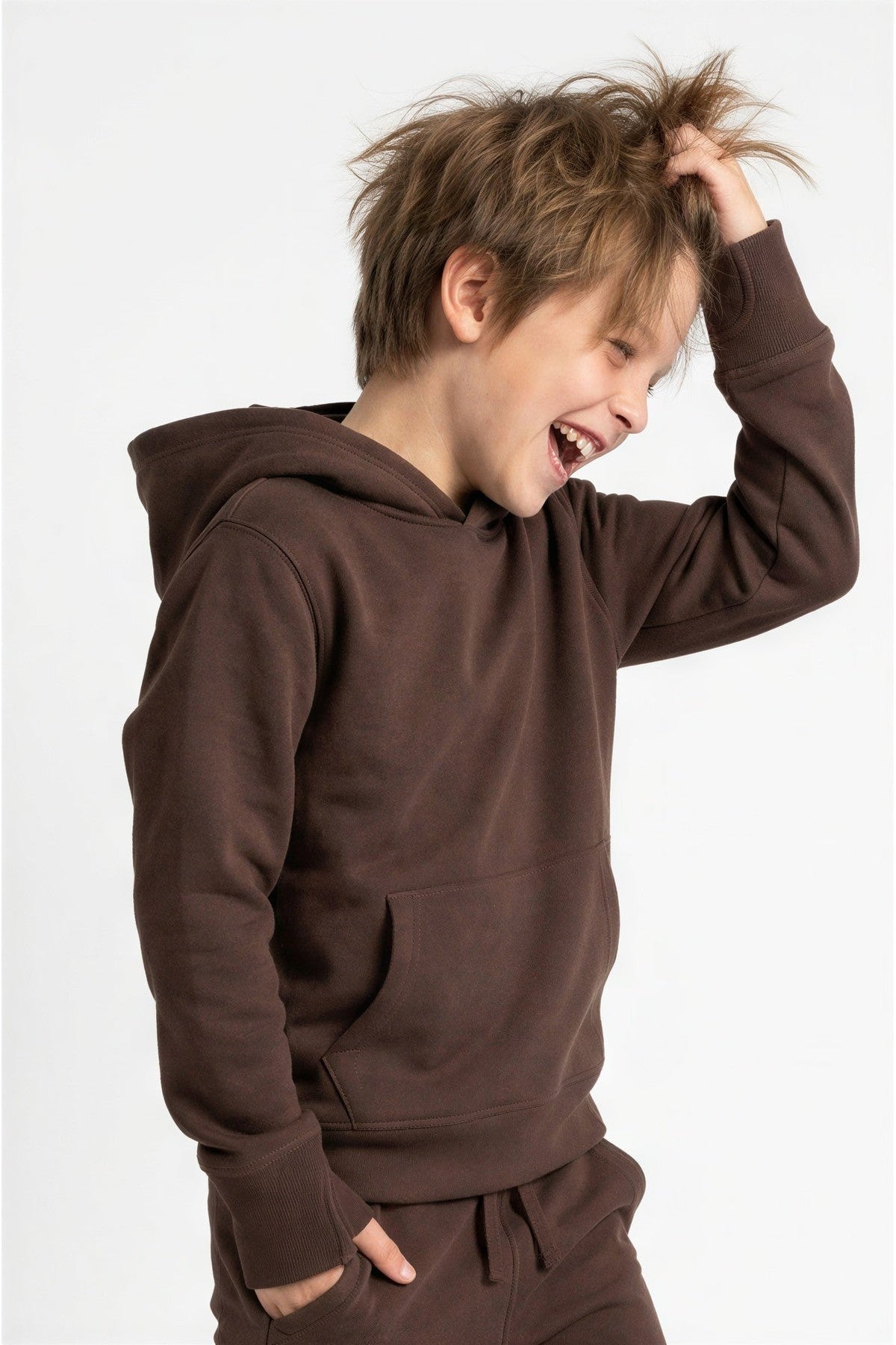 A young boy laughing and looking down while wearing a dark brown pullover hoodie and matching sweatpants against a plain white background. 