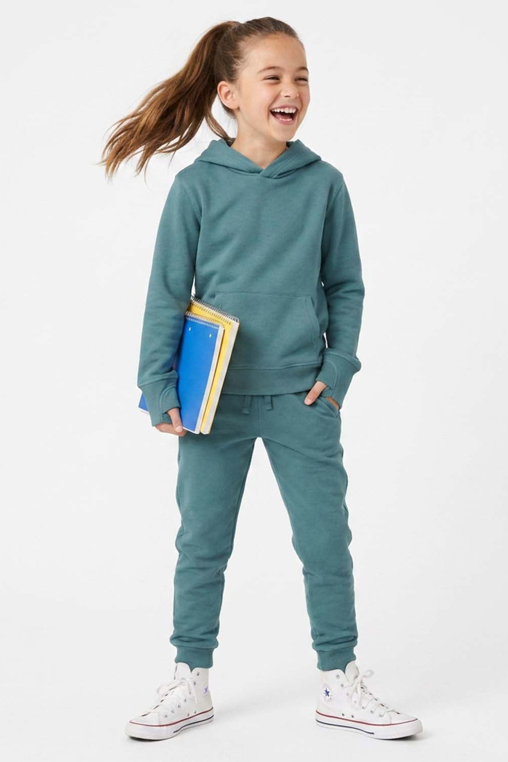 A young girl smiling and laughing while wearing a matching teal green hoodie and sweatpants set, holding a stack of colorful books, and wearing white high-top sneakers against a plain white studio background. 