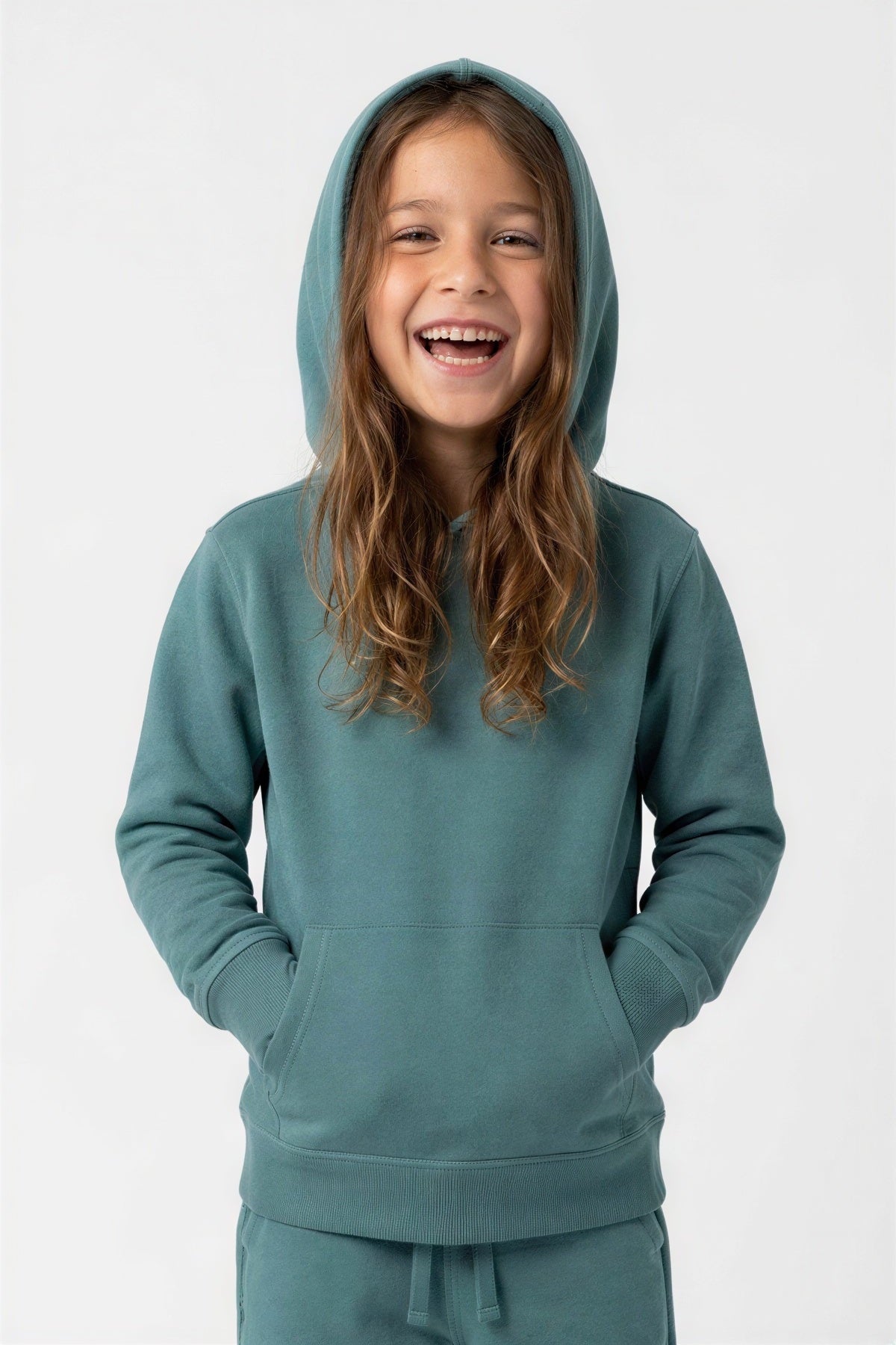 A young girl with long brown hair smiling and laughing while wearing a matching teal blue-green hooded sweatshirt and pants set against a plain light gray background. 