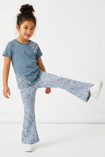 A young girl modeling a matching two-piece pajama set consisting of a slate blue short-sleeve t-shirt with a floral print and matching blue flared pants with a pink and white floral pattern, set against a plain white studio background.