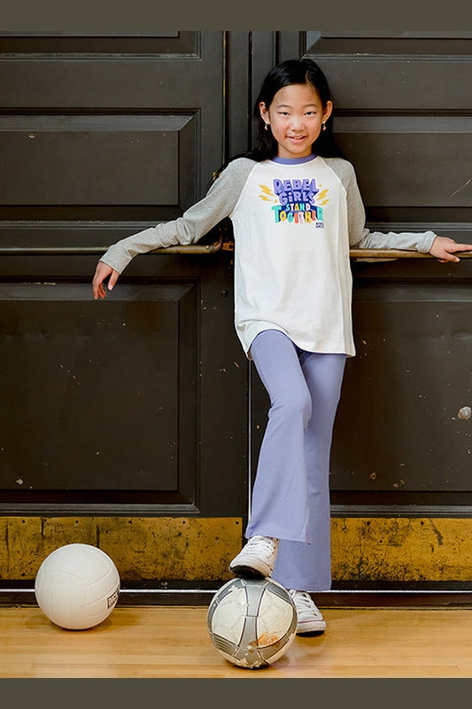 girl wearing a rebel girls graphic long sleeve tee shirt and light purple flared yoga pants with a soccer ball standing against a door