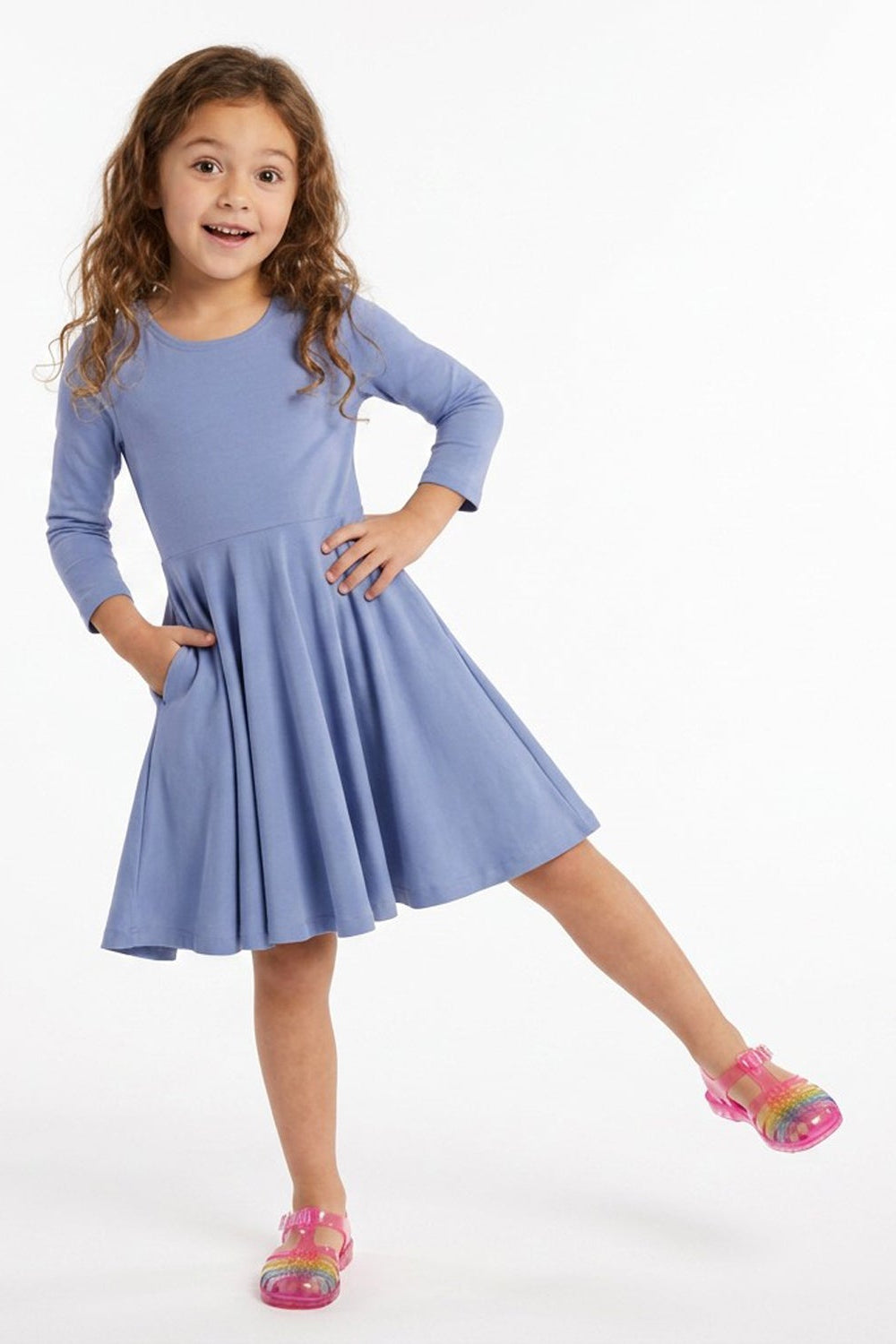 A young girl with curly brown hair wearing a light blue long-sleeve dress with pockets and pink sandals, posing on a white studio background. 