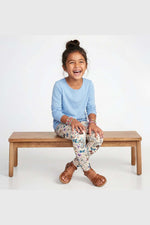 A young girl with her hair in a bun sits smiling on a wooden bench against a white studio background. She is wearing a light blue long-sleeve shirt, colorful floral print leggings, and brown sandals. 