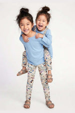 Two young children modeling matching outfits against a plain white studio background. The older child is piggybacking on the younger, and both are wearing light blue long-sleeve tops and floral print leggings. 