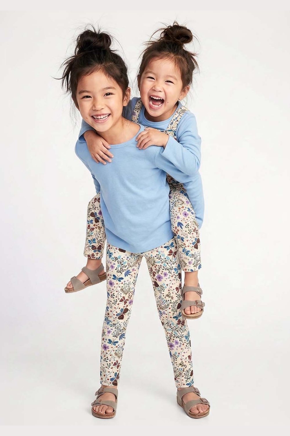 Two young children modeling matching outfits against a plain white studio background. The older child is piggybacking on the younger, and both are wearing light blue long-sleeve tops and floral print leggings. 