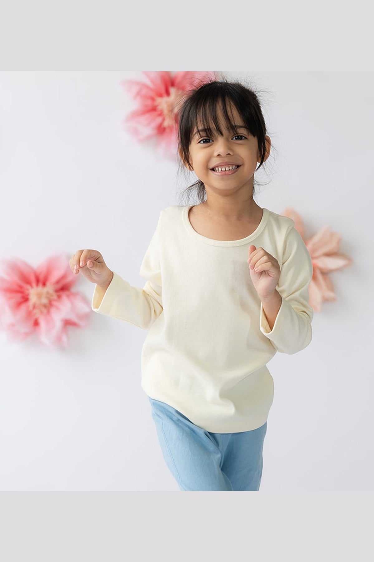 A young girl smiling and posing in a cream-colored long-sleeve crewneck shirt and light blue jeans, set against a white background with soft pink flower decorations. 