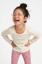 A young girl with her hair in a bun and clear-framed glasses, laughing joyfully while wearing a cream long-sleeve crewneck shirt and pink patterned leggings against a plain white  background. 