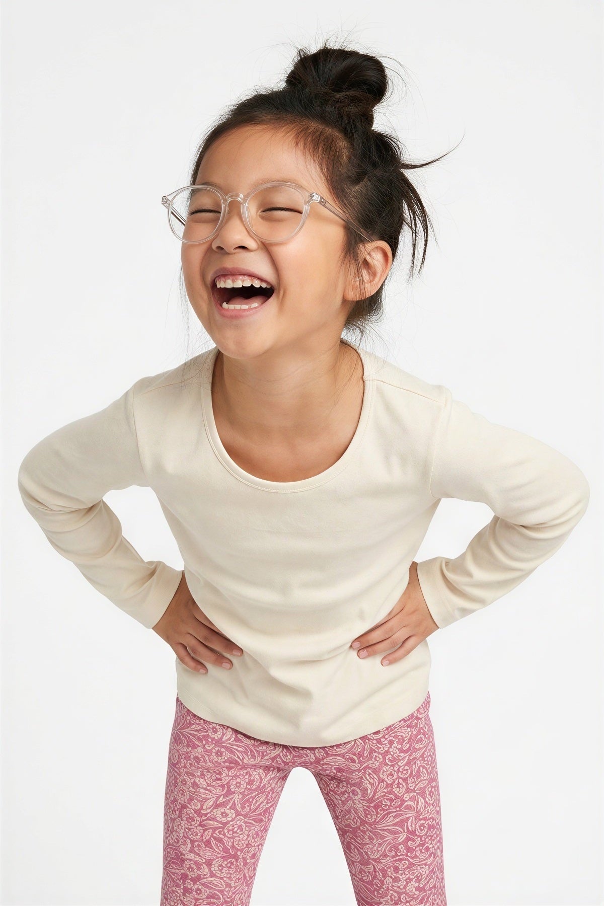 A young girl with her hair in a bun and clear-framed glasses, laughing joyfully while wearing a cream long-sleeve crewneck shirt and pink patterned leggings against a plain white  background. 