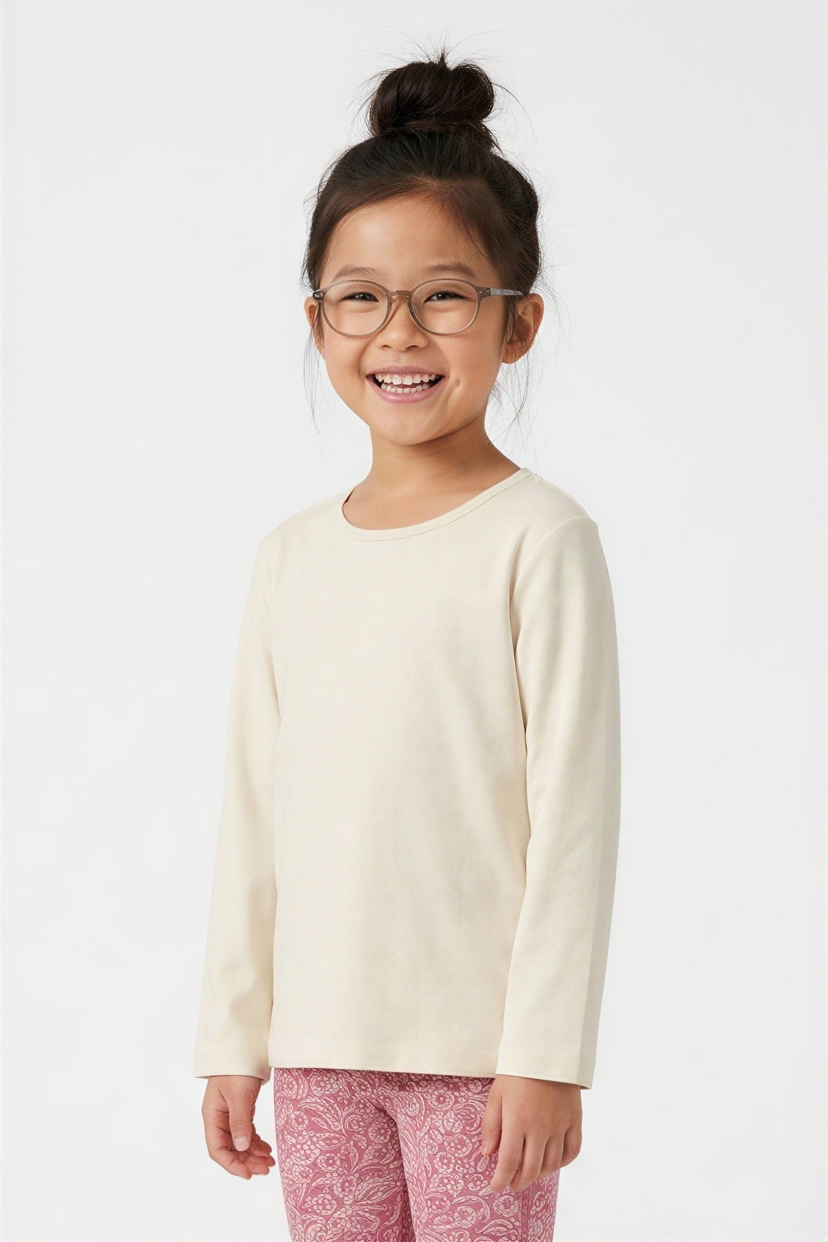 A young girl with glasses and her hair in a bun is smiling, wearing a cream-colored long-sleeve crewneck shirt and pink patterned leggings against a plain white background. 