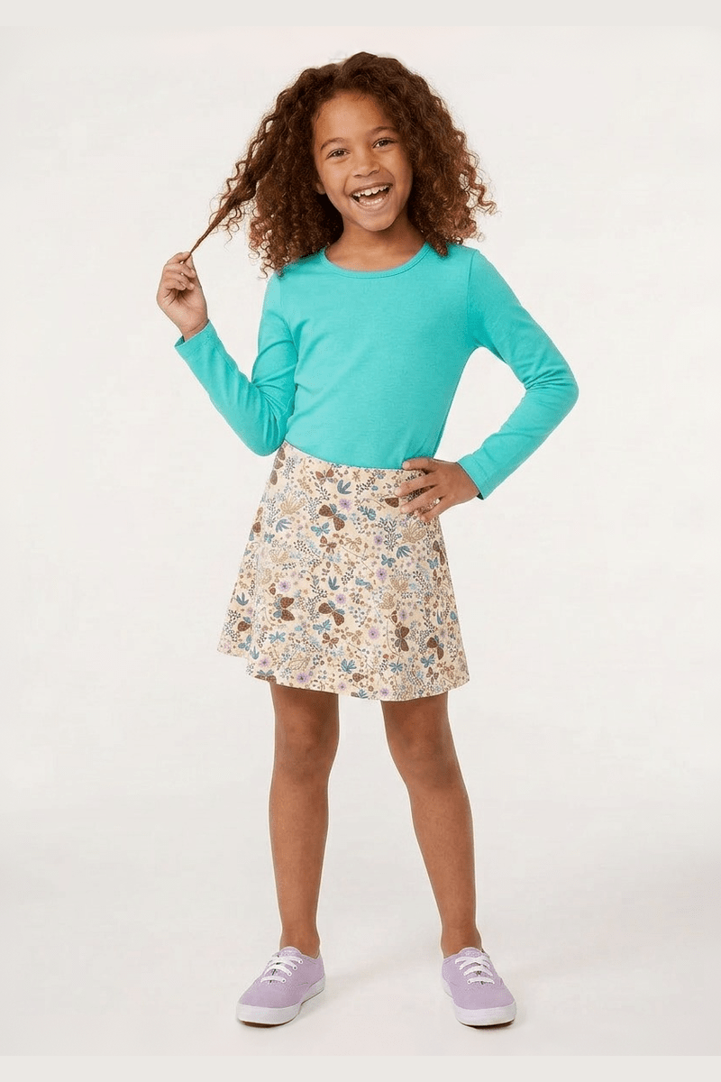 A young girl with curly brown hair stands against a white studio background, wearing a bright turquoise long-sleeve crewneck top and a knee-length A-line skirt with a multicolor floral and bunny print. She is wearing light purple sneakers. 