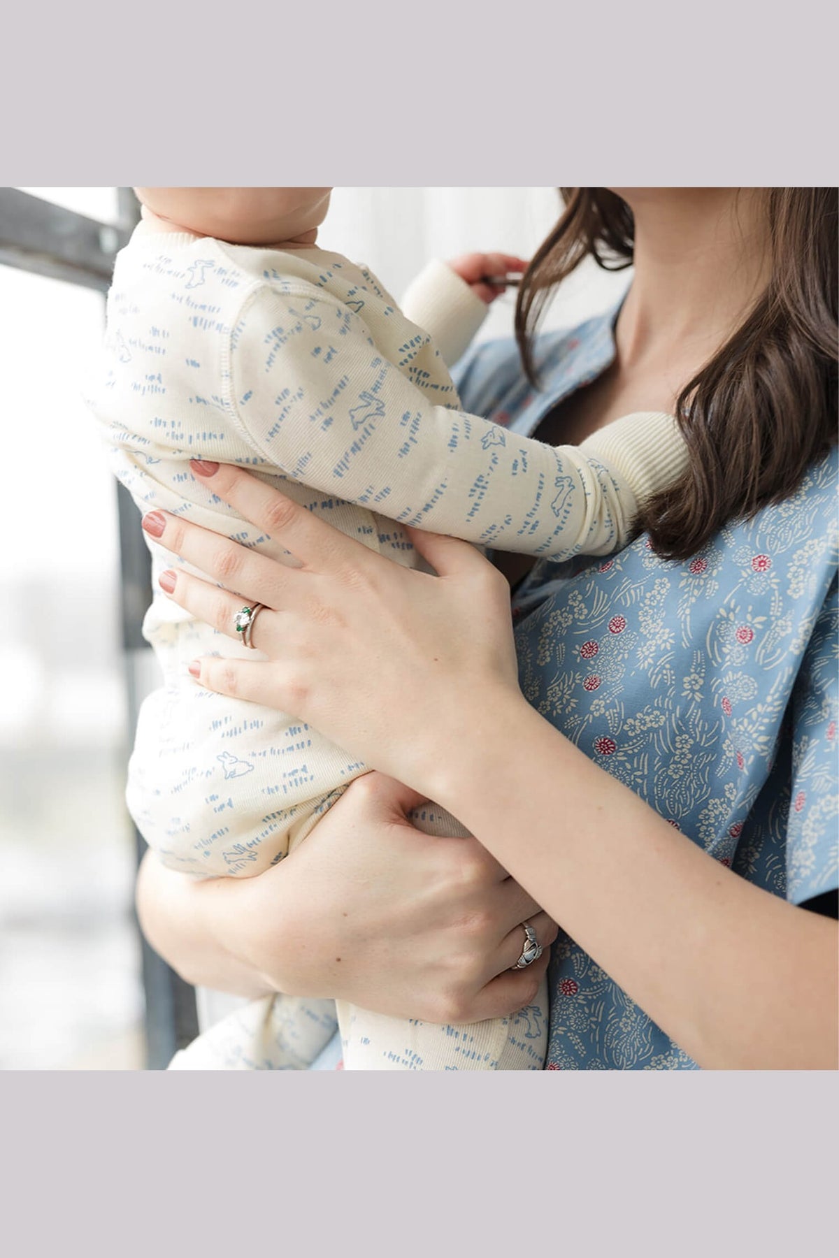 A baby wearing a cream-colored long-sleeve bodysuit with a light blue typographic pattern, being held by a woman wearing a blue floral-patterned top. 