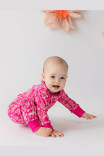 A baby crawling on a white studio floor wearing a bright pink long-sleeve romper with a white floral pattern and solid pink cuffs, with a soft peach flower decoration in the background. 