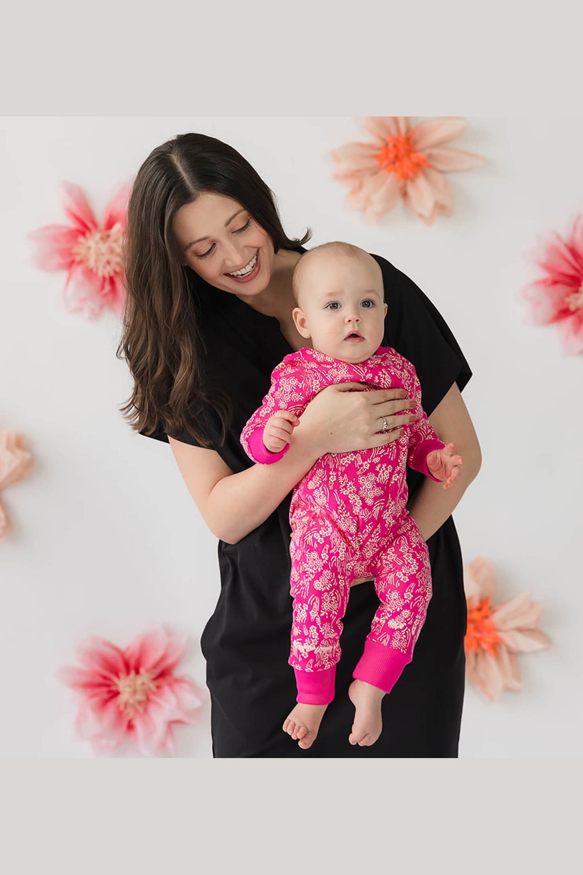 A woman holding a baby wearing a pink floral one-piece pajama set, posed against a white background decorated with large pink and orange flowers. 