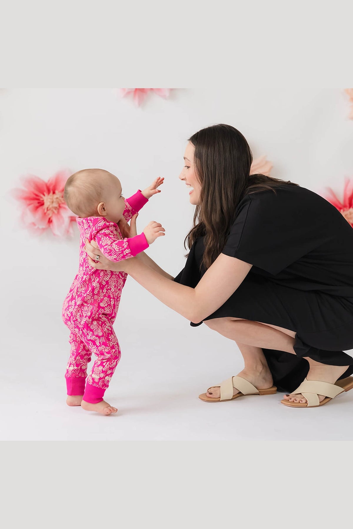 A baby wearing a bright pink floral one-piece pajama stands on a white studio floor while a woman in a black dress crouches to help them touch hands. Pink feather decorations are visible in the background. 