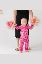 A baby wearing a bright pink floral one-piece jumpsuit stands barefoot while holding hands with an adult wearing a black dress. The background is a clean white studio setting with decorative pink and orange flowers. 