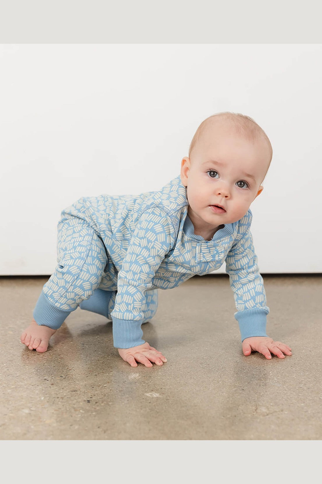 A baby crawling on a concrete floor wearing a light blue and white patterned one-piece jumpsuit. 