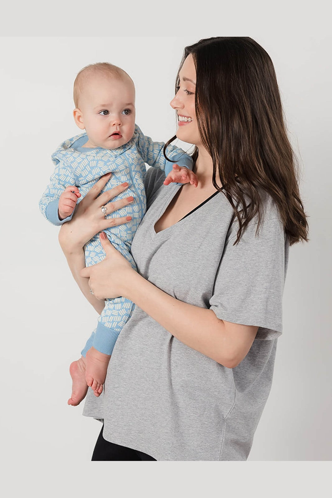 A woman holding a baby wearing a light blue patterned onesie, with the woman wearing a gray V-neck t-shirt, against a plain light gray background. 