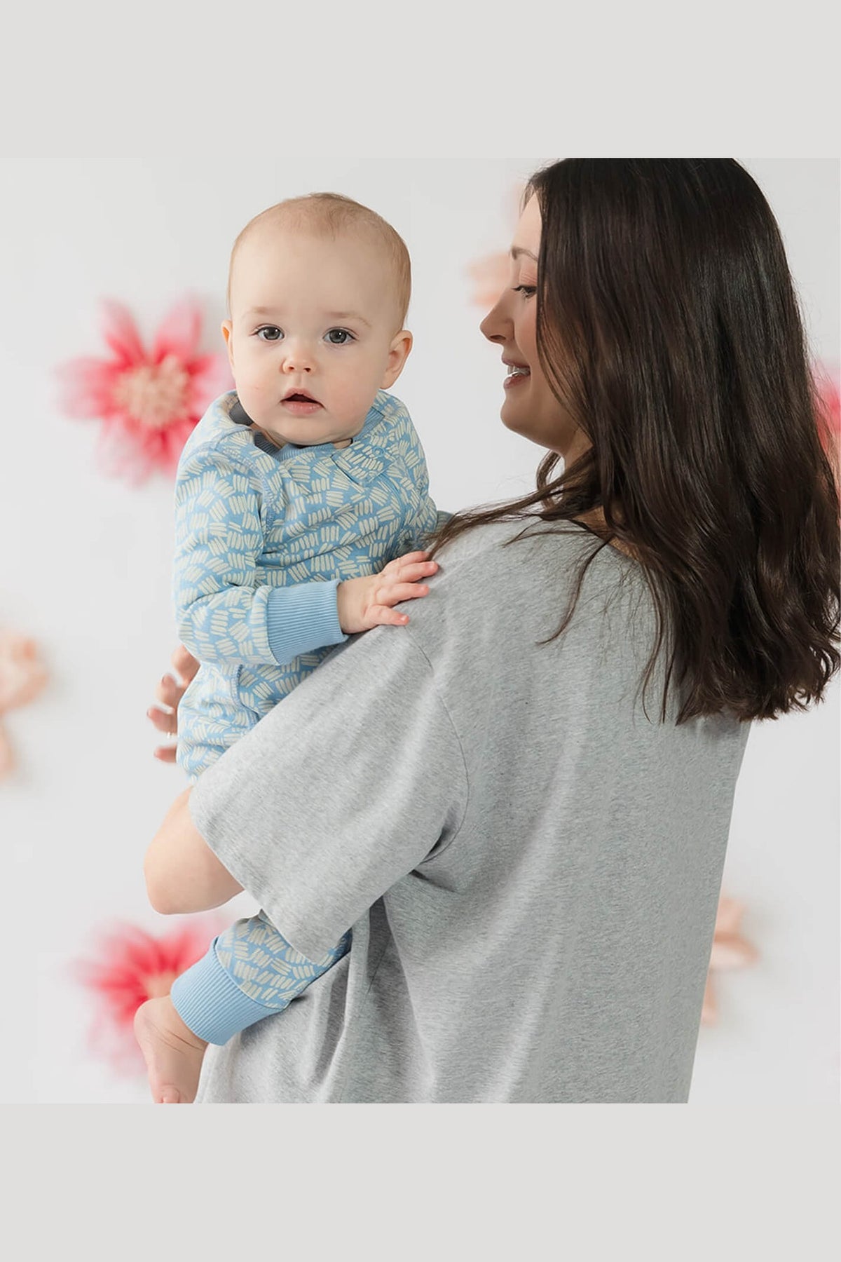 A woman in a light gray oversized top holds a baby wearing a light blue patterned long-sleeve romper. The background is a soft white with blurred pink floral shapes. 