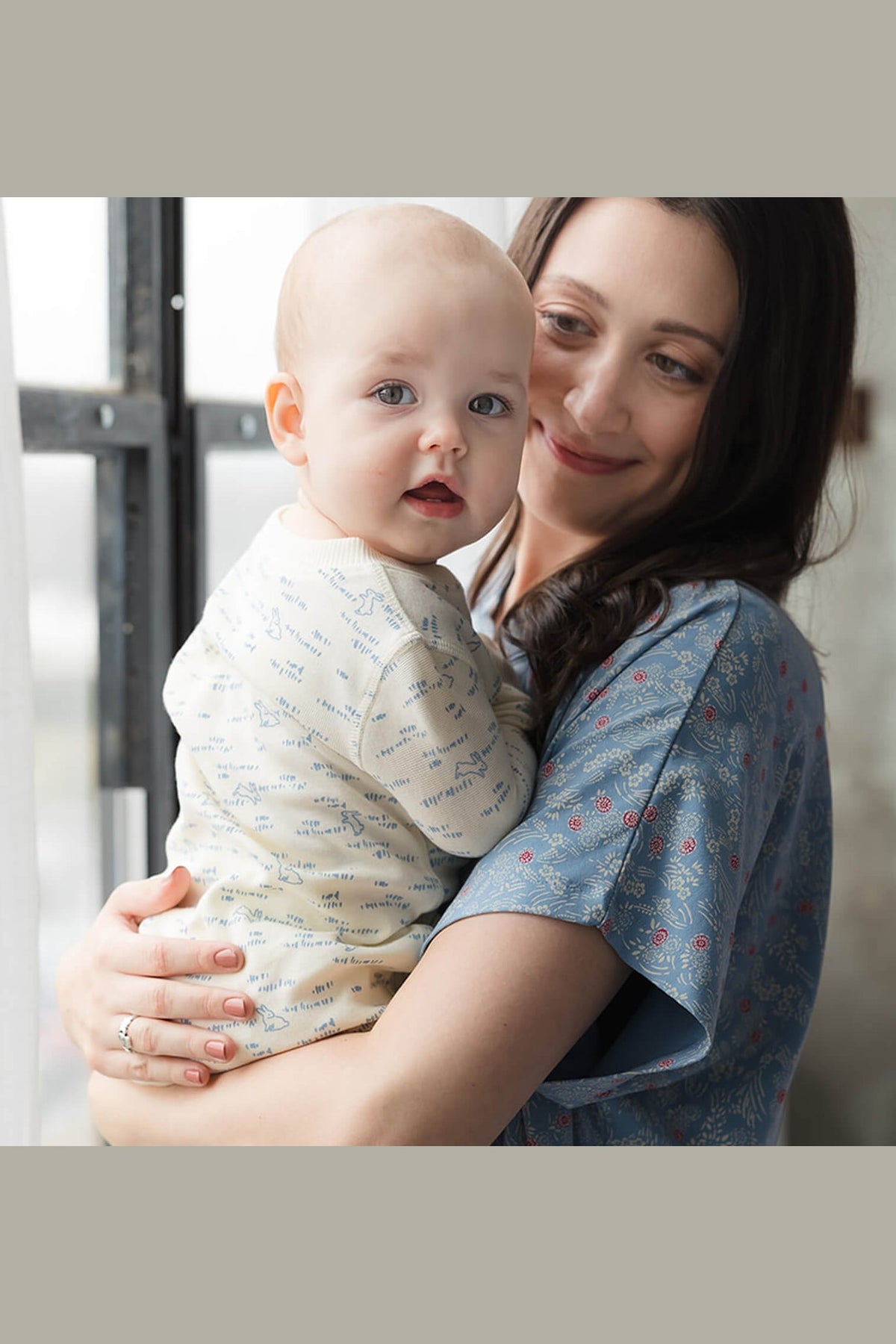 A woman holding a baby wearing a cream-colored patterned onesie, with the woman wearing a blue patterned short-sleeve top. The photo is framed with gray borders on the top and bottom. 