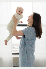 A woman wearing a light blue floral baby wrap or carrier holds a smiling baby dressed in a cream-colored patterned onesie, set against a bright, softly lit window background. 