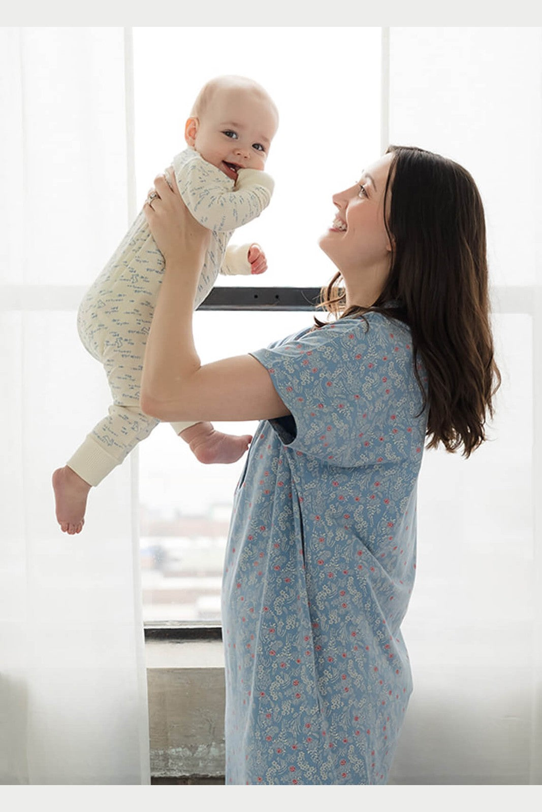 A woman wearing a light blue floral baby wrap or carrier holds a smiling baby dressed in a cream-colored patterned onesie, set against a bright, softly lit window background. 