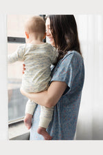 A woman in a blue floral short-sleeve nightgown holds a baby wearing a cream long-sleeve pajama set with a blue pattern, standing by a window. 