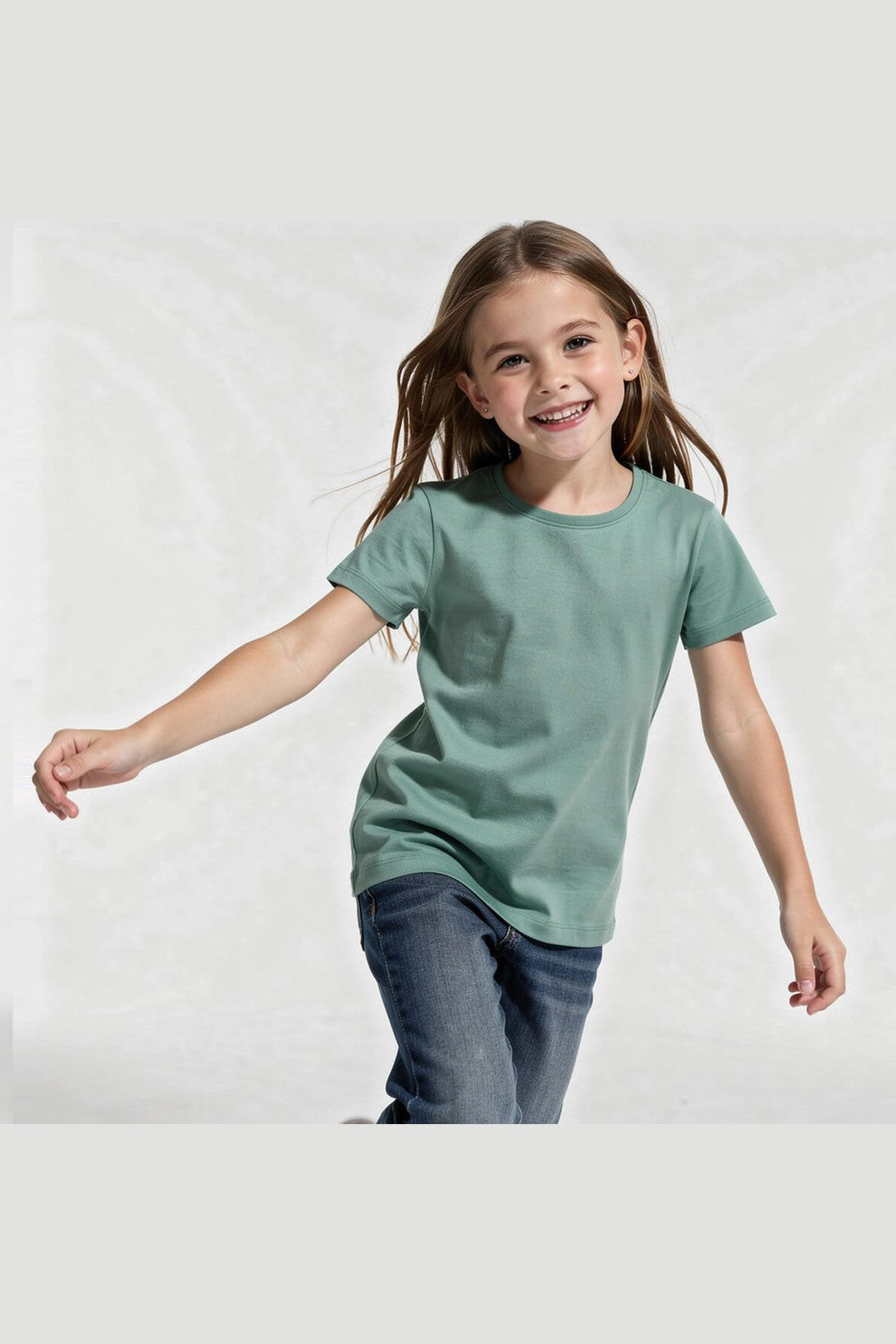 A young girl with long brown hair smiling and running while wearing a sage green short-sleeve crewneck t-shirt and dark blue jeans, set against a light gray studio background. 