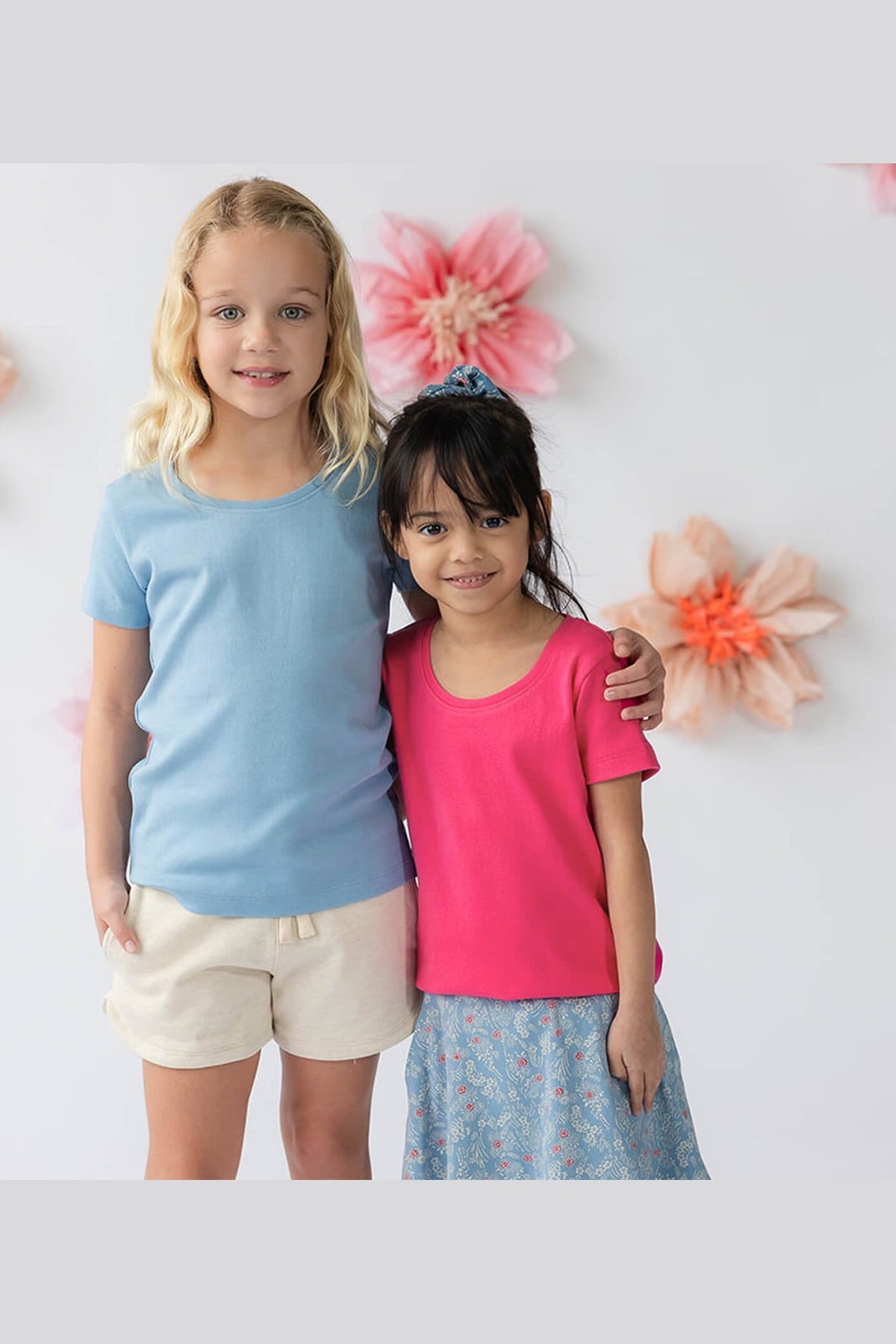 Two young girls standing side-by-side wearing short-sleeve crewneck t-shirts, one in light blue and the other in bright pink, with a white background decorated with paper flowers. 