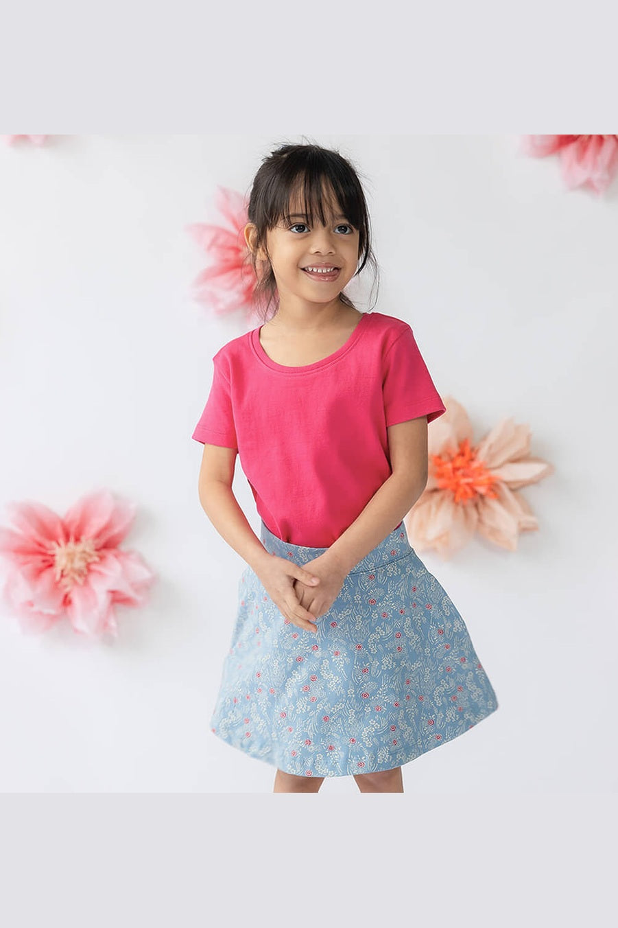 A young girl smiling and posing in a bright pink short-sleeve t-shirt and a light blue floral print skirt, set against a white wall decorated with soft pink paper flowers. 