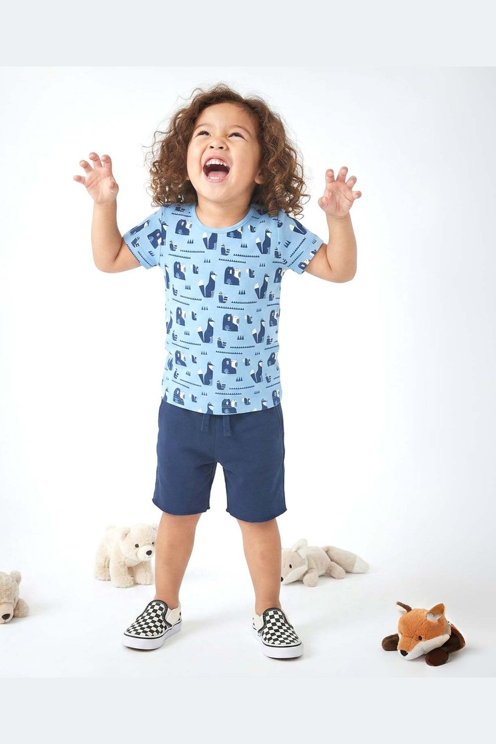 Child wearing a blue patterned shirt and navy shorts with teddy bears in the background 