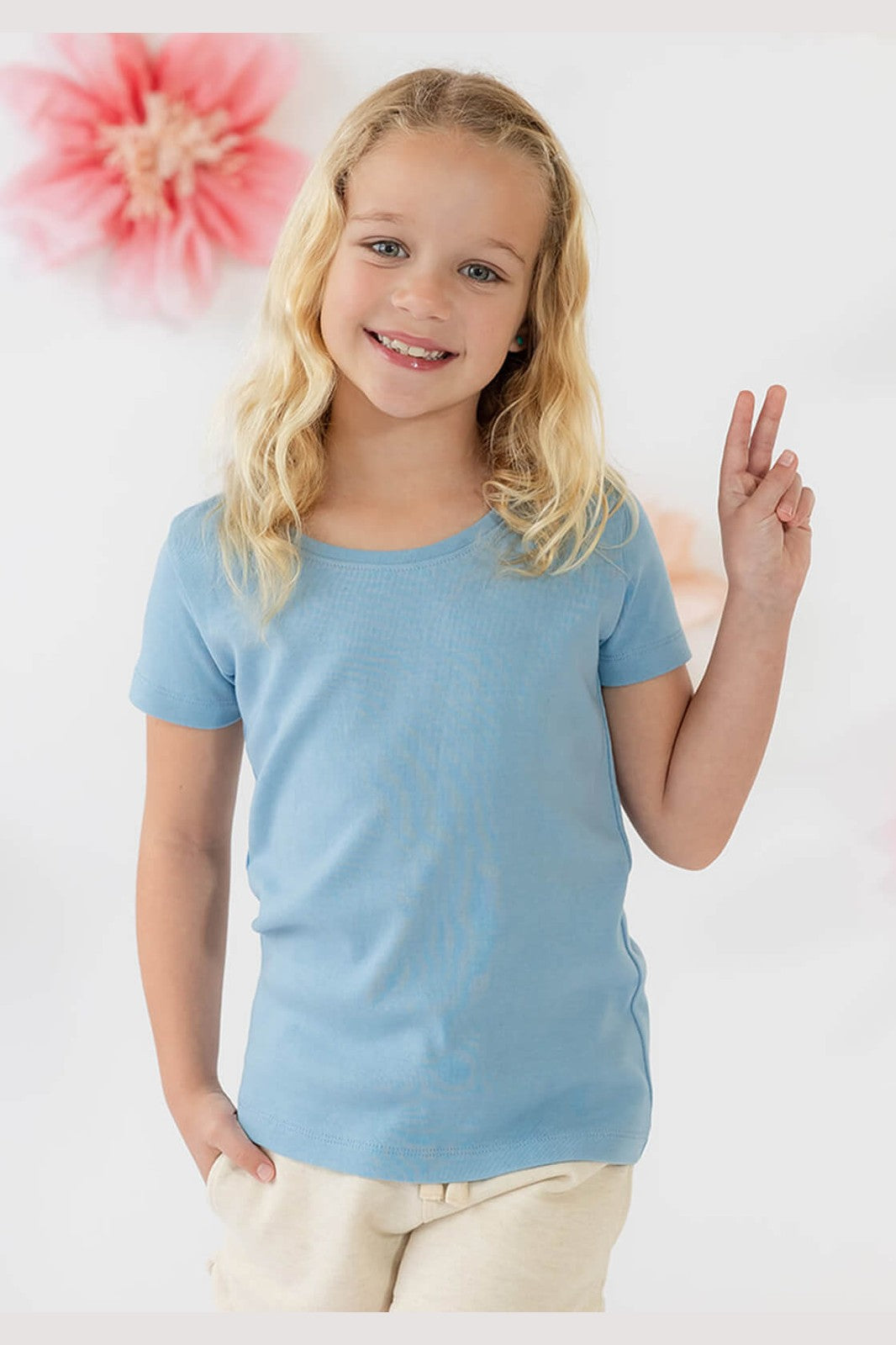 A young girl with blonde hair smiling and posing, wearing a light blue short-sleeve crewneck t-shirt and cream pants against a white background with a pink flower decoration. 