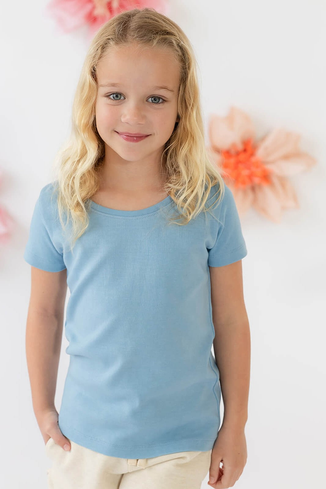 A young girl with blonde hair wearing a light blue short-sleeve crewneck t-shirt and cream pants, standing against a white wall with soft pink and orange flower decorations. 