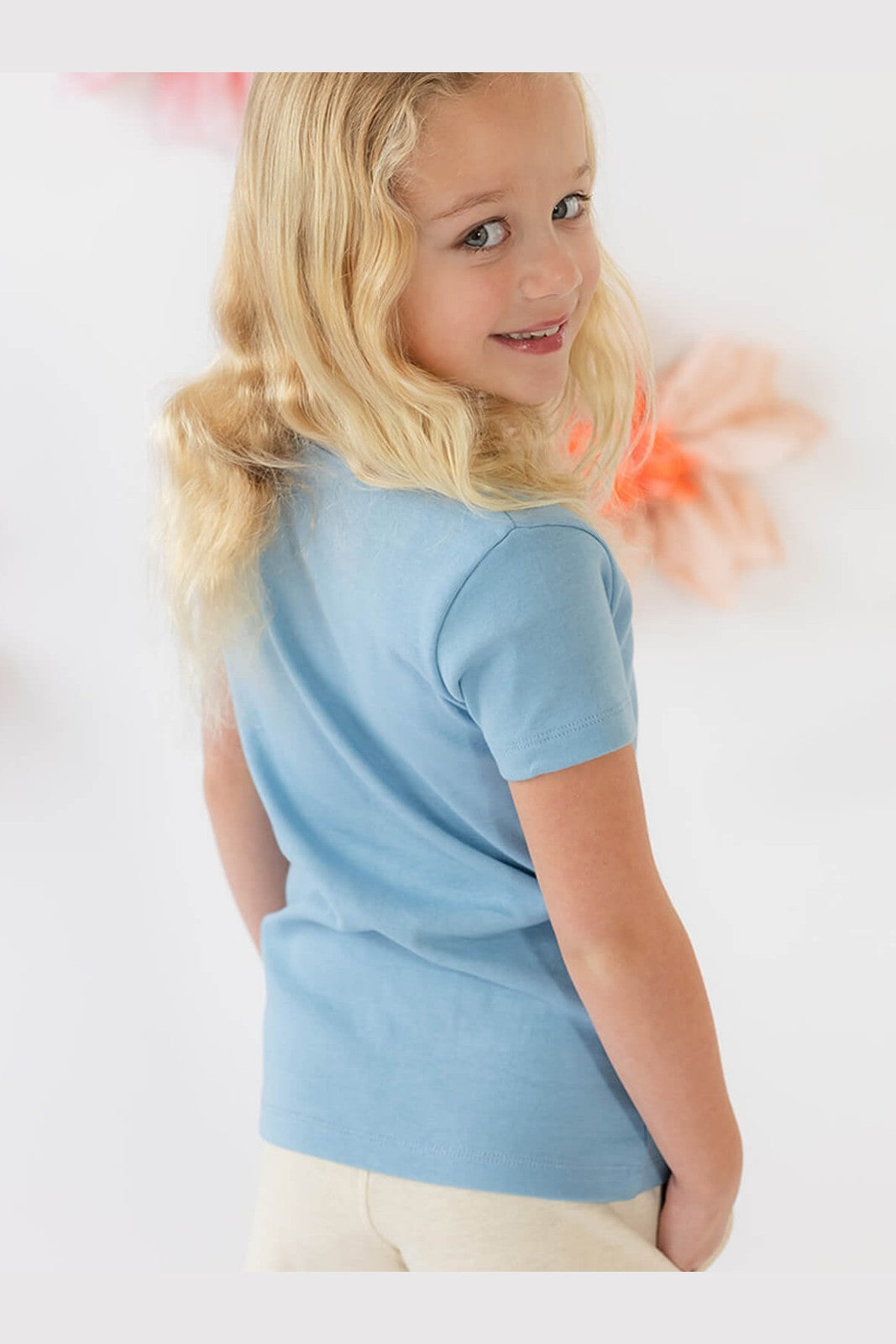 A young blonde girl wearing a light blue short-sleeve t-shirt and cream pants, shown from a side-back angle against a bright white background with soft pink decorative elements. 